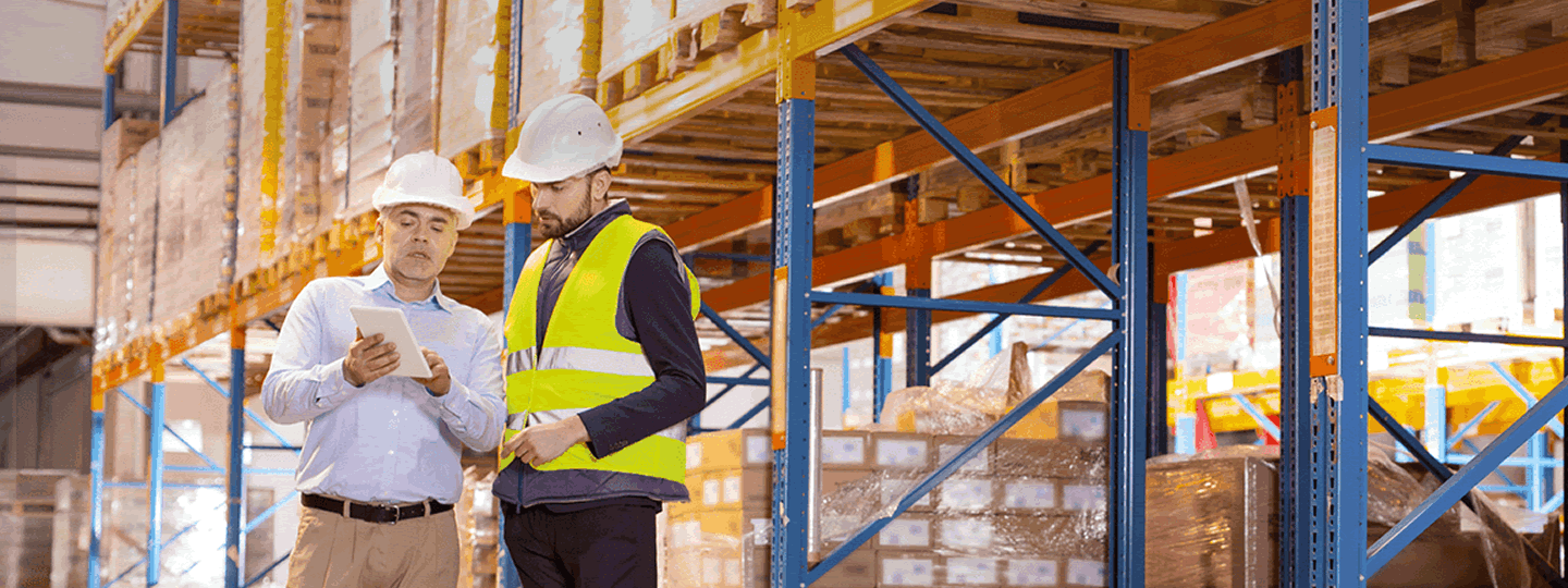 Two workers in safety helmets and vests stand in a warehouse, looking at a tablet. Shelves filled with boxes and packages are visible in the background.