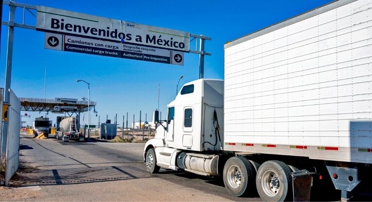 A white semi-truck approaches a border crossing under a large sign reading Bienvenidos a México on a clear, sunny day. The sign indicates directions for commercial cargo trucks.