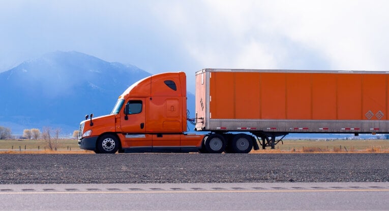 A bright orange semi-truck and trailer parked on a paved road, with open fields and mountains in the background under a cloudy sky.