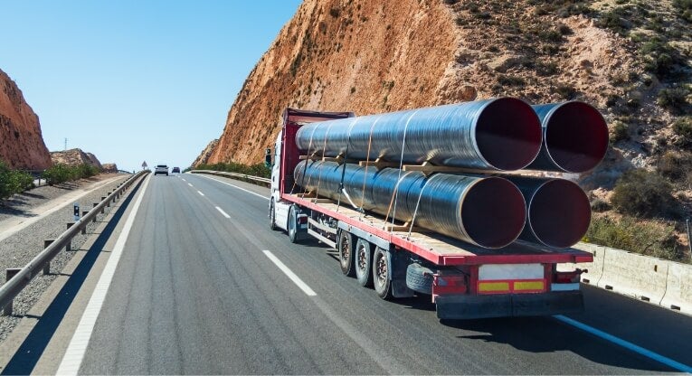 A flatbed truck transports large metal pipes on a highway passing through rocky hills under a clear blue sky.