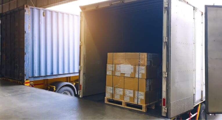 A shipping truck backed up to a loading dock with its door open, showing a pallet stacked with cardboard boxes inside. Another shipping container is visible in the background.