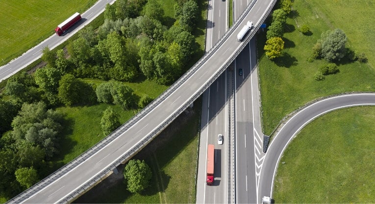 Aerial view of a highway interchange surrounded by green trees and grass, with several trucks and cars traveling on the roads and overpasses.