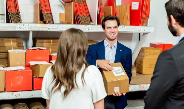 A man in a blue suit holds a package and talks to two people in a warehouse, with shelves of boxes and packages in the background, showcasing UPS and Worldwide Express services.