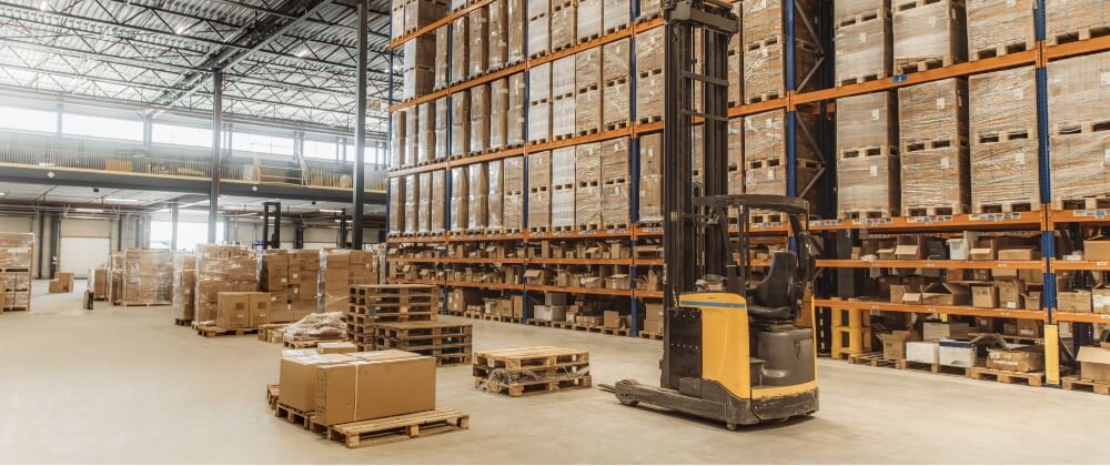A spacious warehouse with tall shelves stacked with boxes and pallets. A yellow forklift is parked near some stacked pallets and cardboard boxes on the warehouse floor.