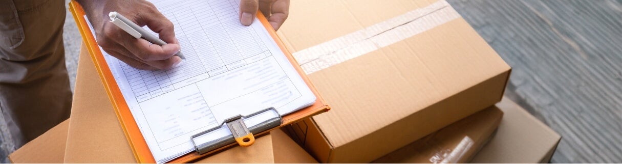 A person holding a pen writes on a clipboard with a form or checklist, next to several stacked cardboard boxes, suggesting a delivery or inventory process.