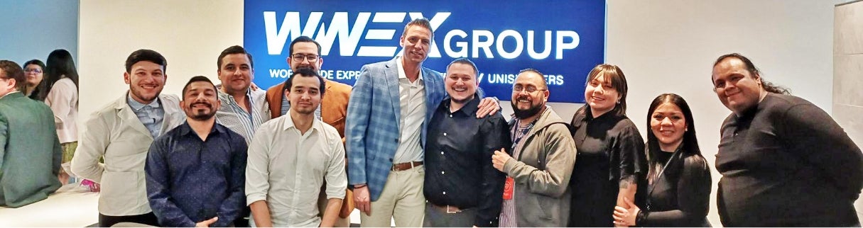 A group of twelve people stand smiling in front of a blue sign that reads WWEX GROUP. They appear to be colleagues specializing in cross border freight gathered in a professional office setting.