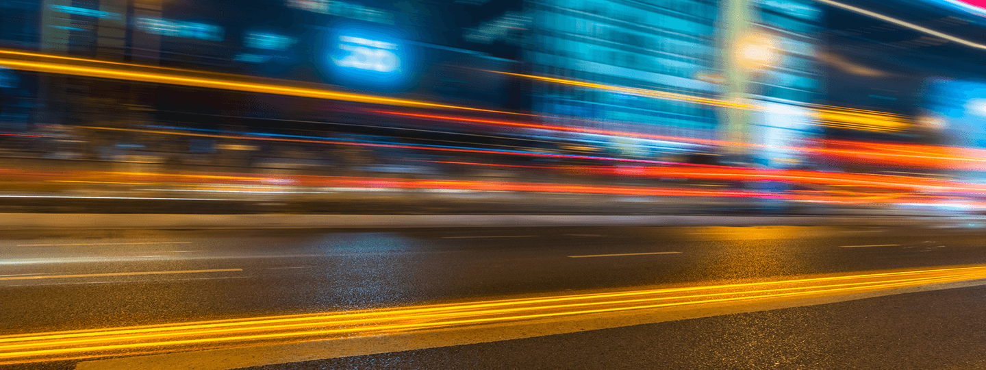 A city street at night with bright, colorful light trails from passing vehicles, creating a sense of fast movement and energy. Blurred buildings and lights are visible in the background.