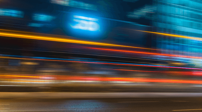 Blurred city street at night with colorful streaks of light from moving vehicles and illuminated buildings, creating a sense of motion and speed.
