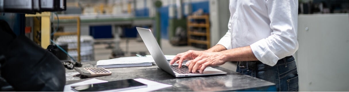 A person in a white shirt uses a laptop on a metal workbench in an industrial or factory setting. Nearby are a calculator, notebook, and smartphone. The background shows shelves and machinery.