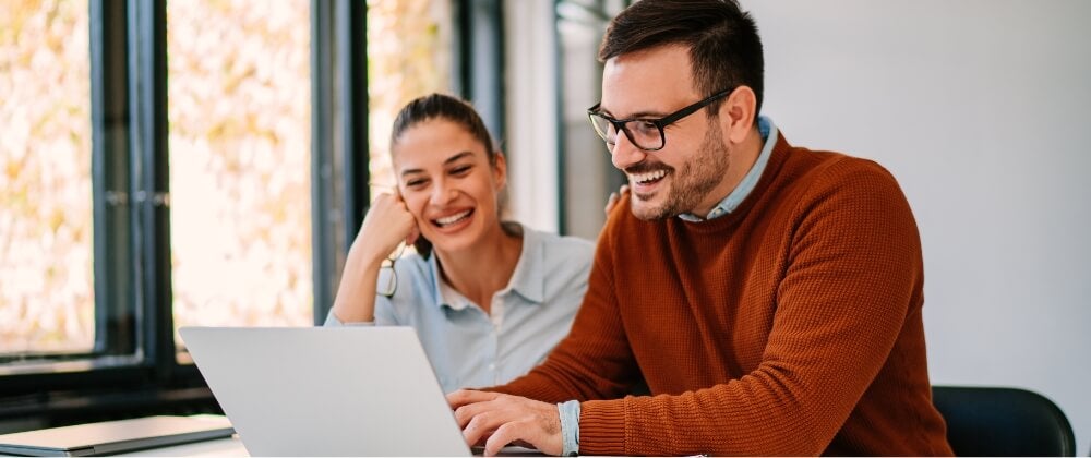 Two people sit at a desk, smiling and looking at a laptop screen. The man wears glasses and a rust-colored sweater, while the woman wears a light blue shirt. They appear to be working together in a bright office space.