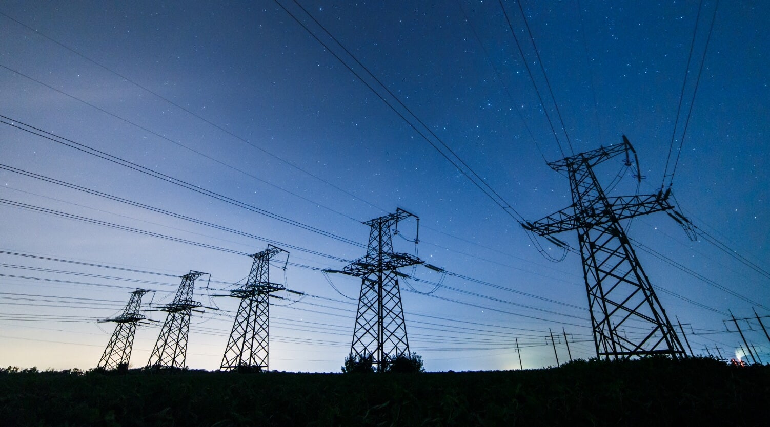 High-voltage power lines and tall electrical towers stretch across a field at dusk, with a clear sky and stars visible in the background.