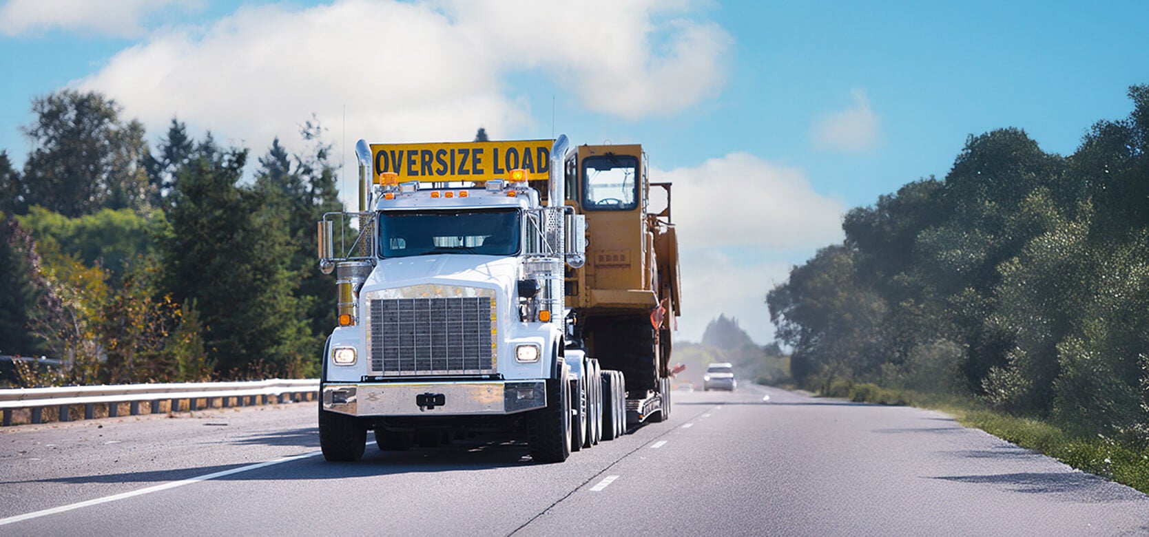 A large white truck carrying construction equipment with an OVERSIZE LOAD sign drives down a divided highway on a clear day, surrounded by trees and a few distant vehicles.
