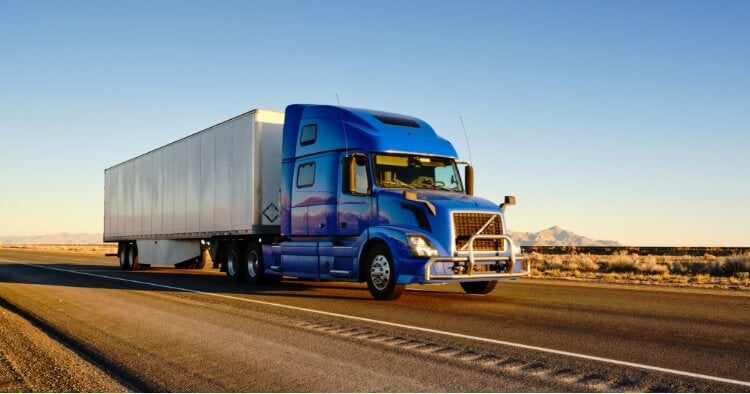 A blue semi-truck with a white trailer drives on a highway through a flat, dry landscape under a clear blue sky, with mountains visible in the distance.