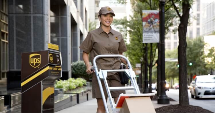 A UPS delivery person in uniform smiles while pushing a hand truck with packages on a city sidewalk, near a UPS drop box and office buildings.
