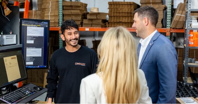 Three people stand and converse in a warehouse office. One man in a black shirt smiles at a man in a blue suit, while a woman with long blonde hair stands with her back to the camera. Monitors and paperwork are visible.