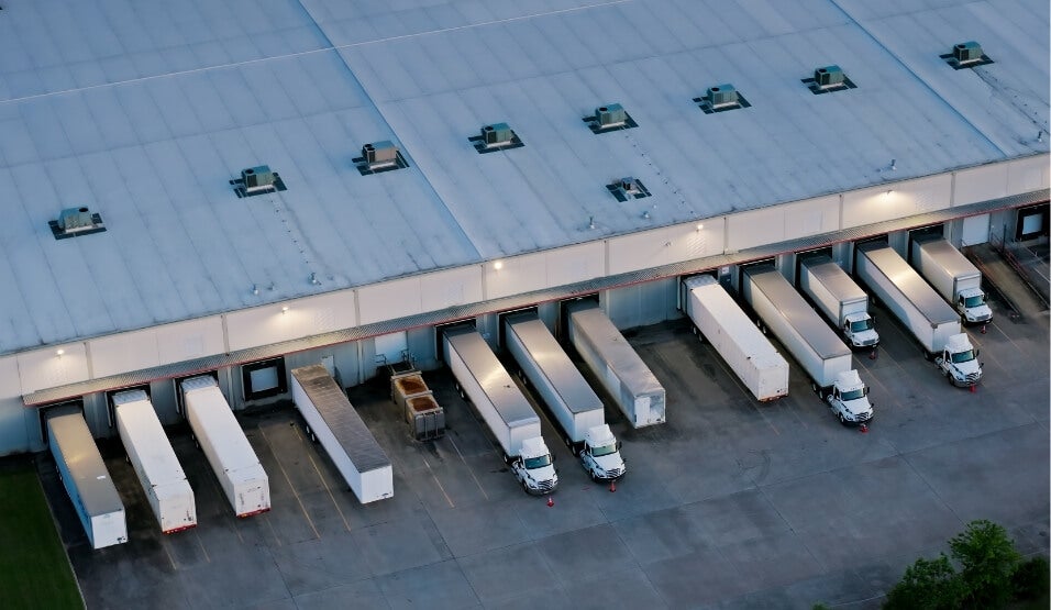 Aerial view of loading docks at a large warehouse, with eight white semi-trucks backed into separate dock doors, ready for loading or unloading. The area is well-lit, and the concrete lot is mostly empty.