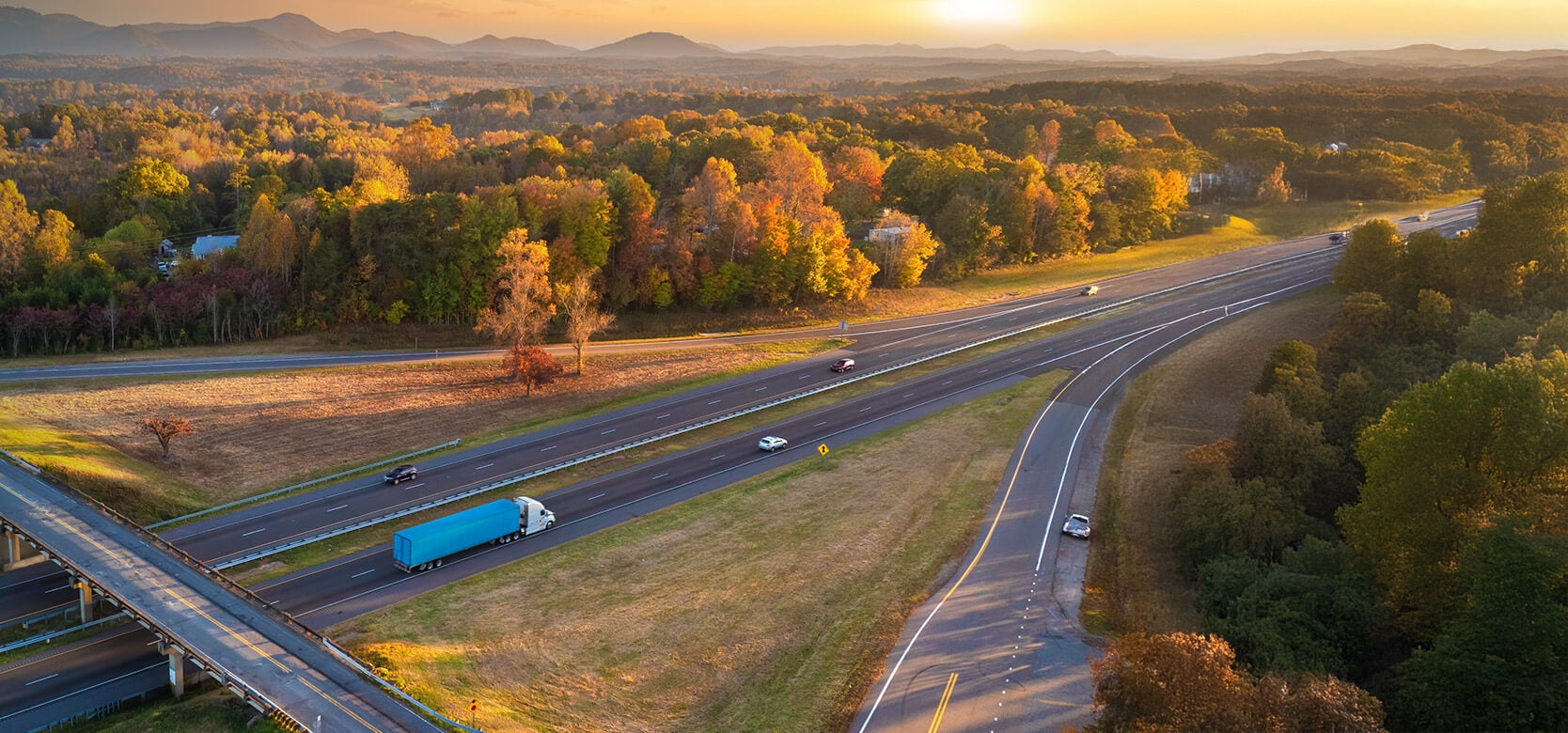 Aerial view of a highway with cars and a blue truck driving through a forested area with autumn foliage; mountains are visible in the background at sunset.