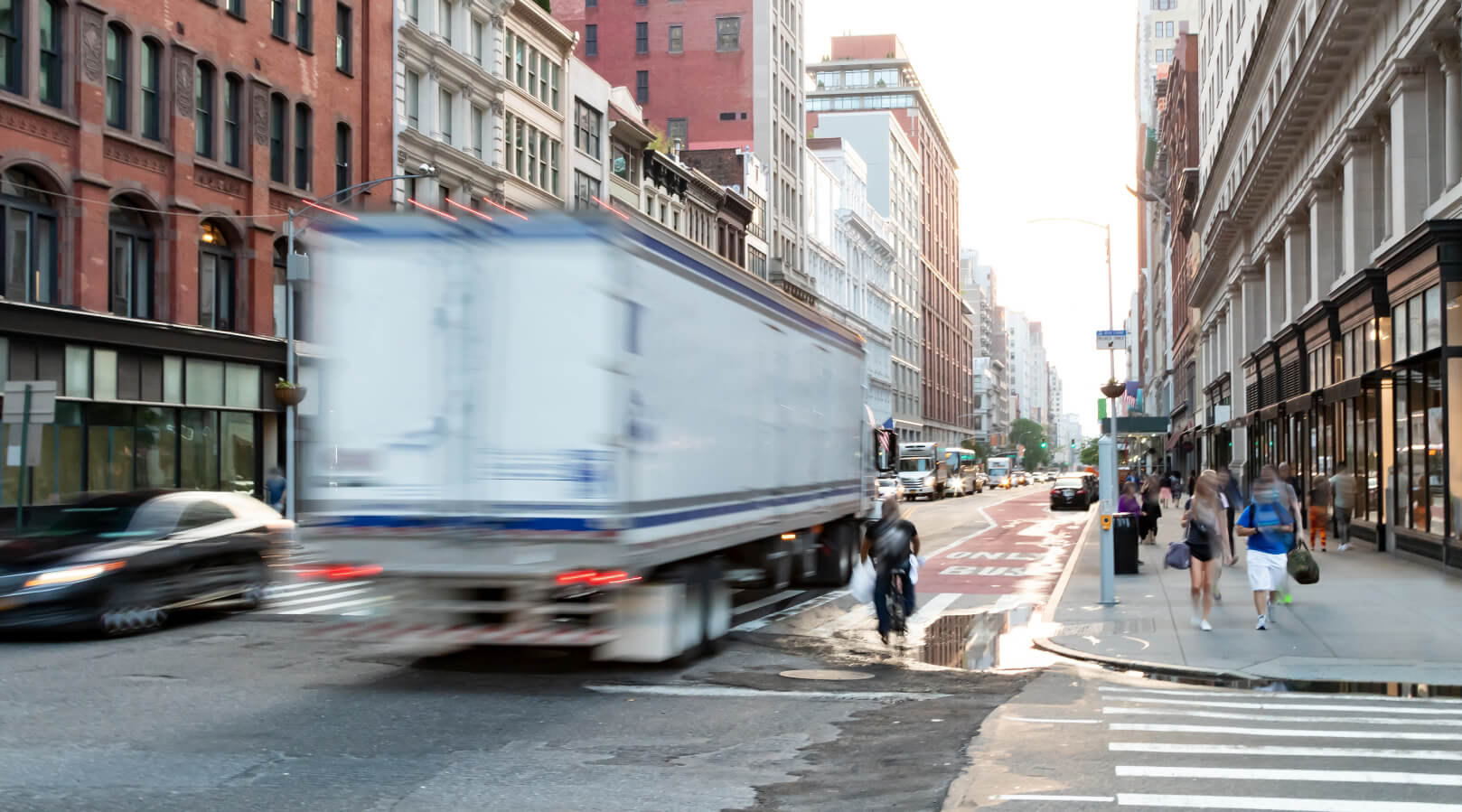 A blurry white truck speeds through a city street lined with tall buildings, shops, and pedestrians on the sidewalk during daylight. Other cars and people are visible in the background.