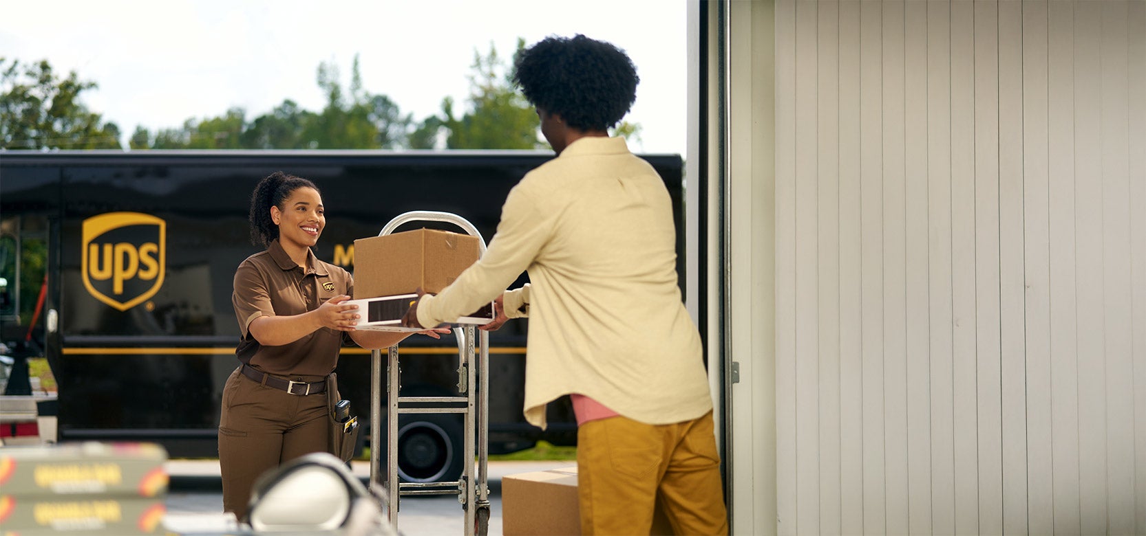 A UPS delivery driver hands a package to a person at an open doorway, with a UPS truck parked outside in the background.