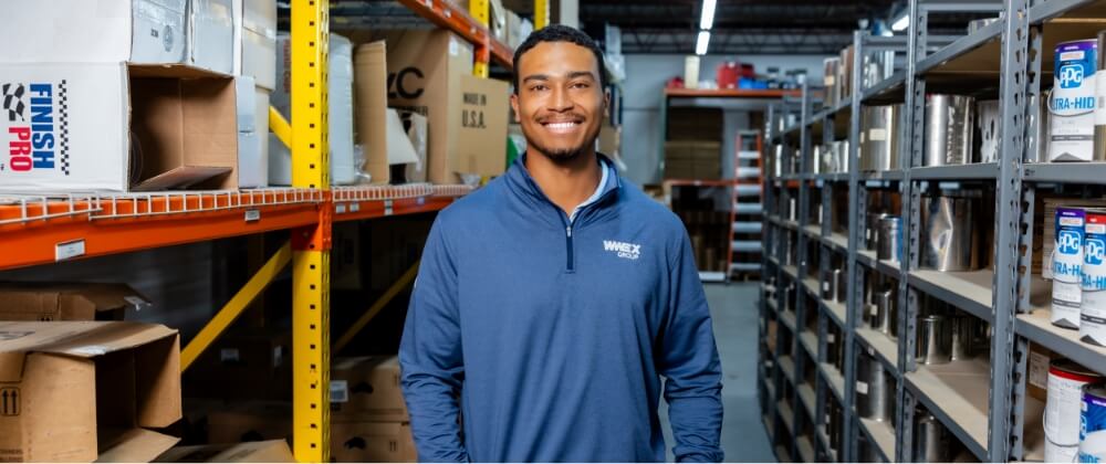 A man in a blue long-sleeve shirt stands smiling in a warehouse aisle surrounded by metal shelves filled with boxes and containers. Bright lighting highlights the organized industrial setting.