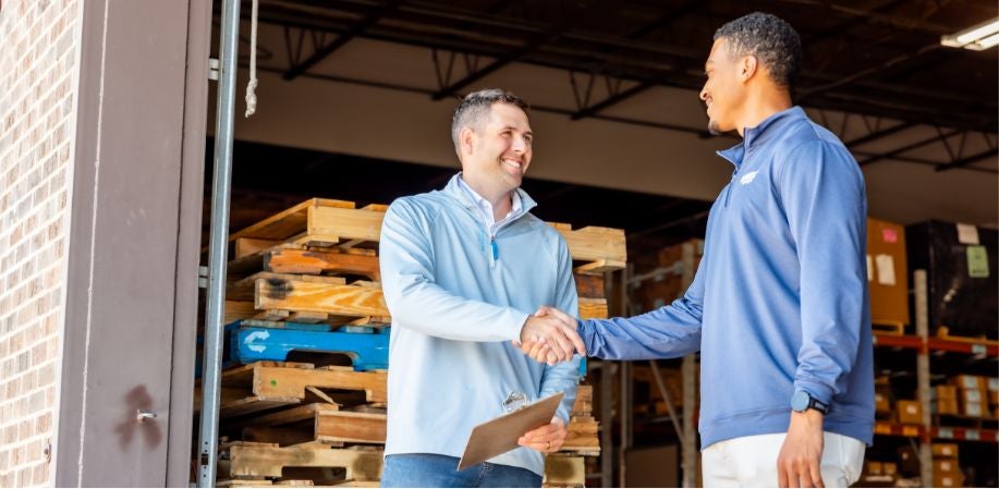 Two men stand outside a warehouse shacking hands.
