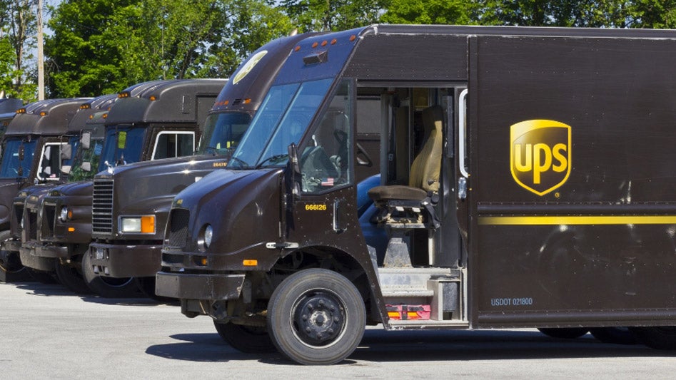 Several brown UPS and Worldwide Express delivery trucks are parked in a row in a lot, with trees visible in the background. The door of the nearest truck is open, revealing the driver's seat inside.