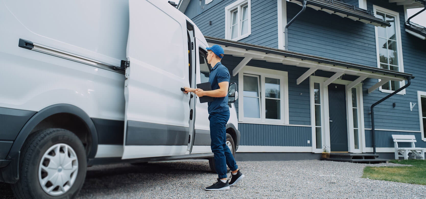 A delivery person in a blue uniform and cap stands by an open white van, holding a package, outside a modern blue house with white trim on a gravel driveway.