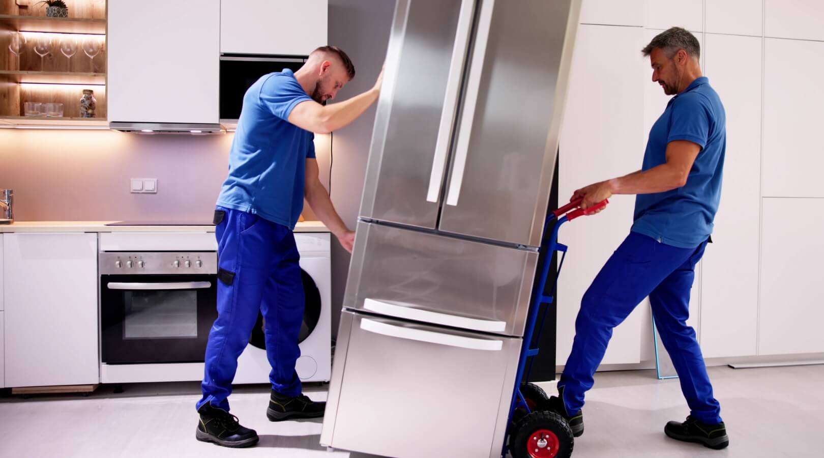 Two movers in blue uniforms are relocating a large stainless steel refrigerator in a modern white kitchen. One man steadies the fridge while the other uses a hand truck to move it.