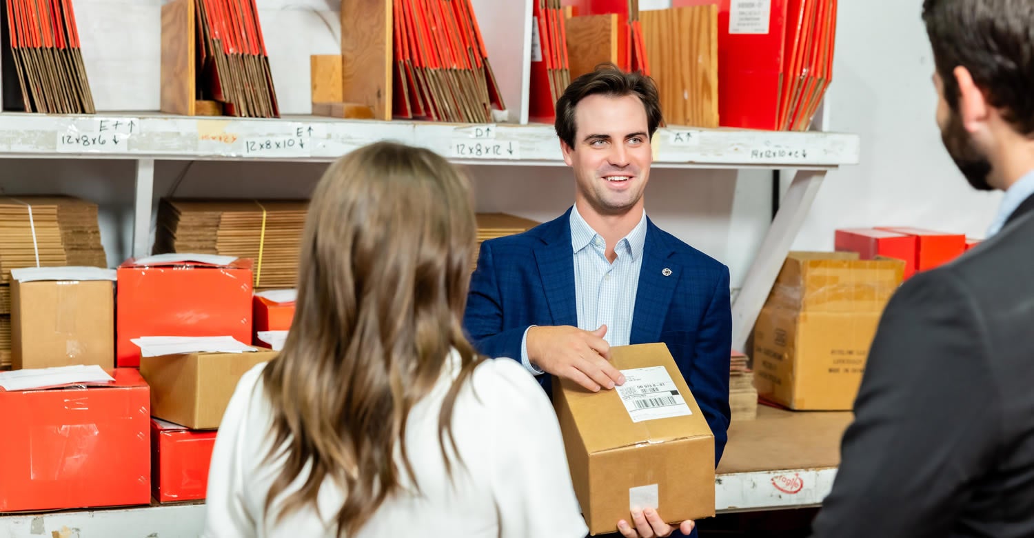 A man in a blue suit holds a cardboard box and smiles while talking to two people in a storage room filled with packages and shipping supplies.
