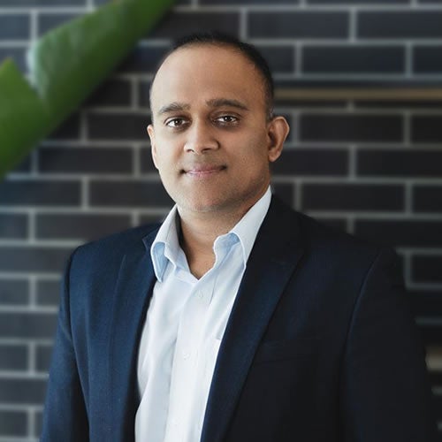 A man, possibly Arjun Srinivasan, wearing a dark suit and white dress shirt stands in front of a dark brick wall, looking at the camera with a neutral expression. A large green leaf appears in the upper left corner, hinting at an xModal setting.