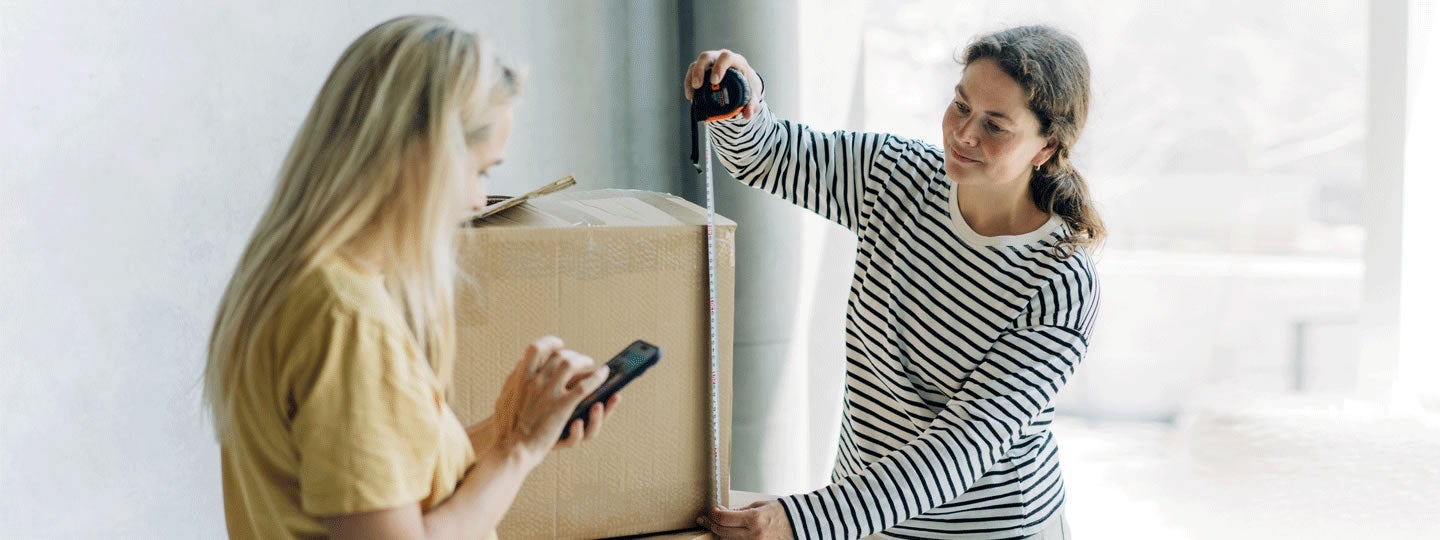 Two women are preparing a cardboard box; one measures it while the other checks a smartphone. They are working together in a bright indoor space, using AI in logistics to streamline their shipping process.