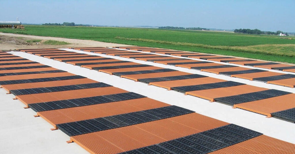 Several rows of orange and black rectangular slatted panels, prepared for emergency shipping by Greater Good Charities, are arranged on a large concrete surface in an open rural area with fields and a clear sky in the background.
