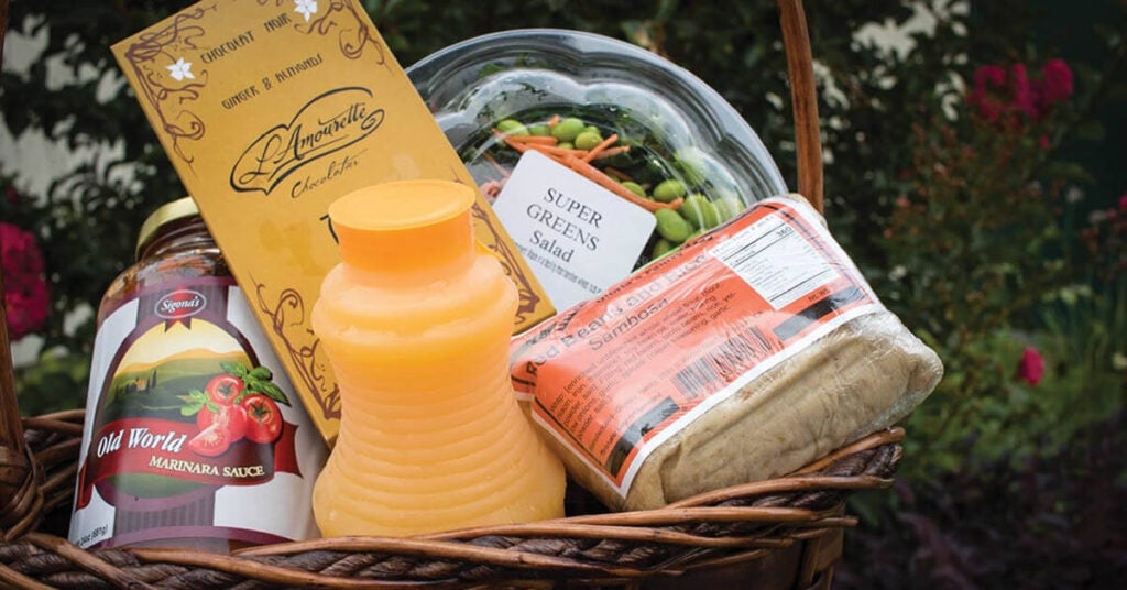 A wicker basket filled with marinara sauce, orange juice, chocolate, super greens salad, and packaged cheese—set outdoors among greenery and flowers—showcases ADA Enterprises’ commitment to quality in B2B logistics.