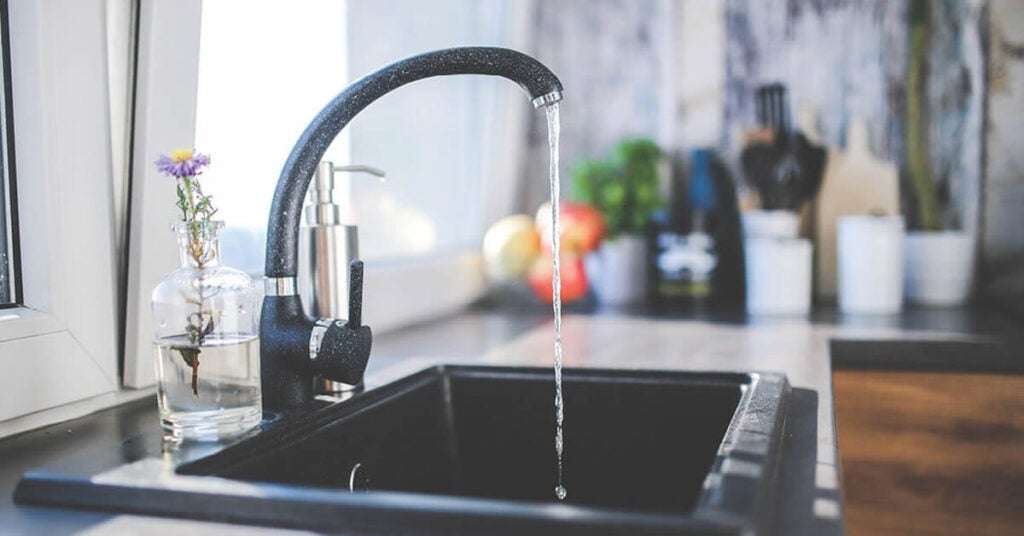 A modern black kitchen faucet with water running into a black sink. A soap dispenser and a small glass vase with wildflowers sit on the counter, while hints of kitchen utensils, fruit, and temperature-controlled shipping boxes blur in the background.