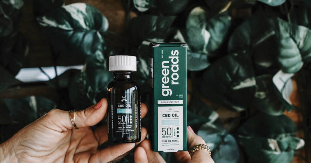 A person holds a bottle of CBD oil and its box labeled green roads in front of leafy green plants. The label shows 50mg per serving and 1500mg total broad spectrum CBD oil, highlighting the importance of efficient manufacturing logistics.