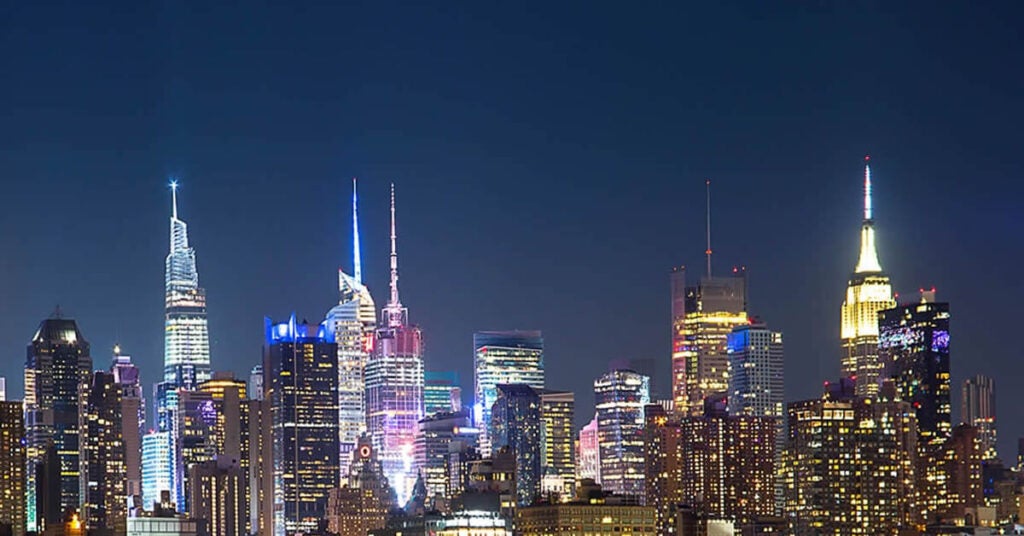 A brightly lit city skyline at night features tall skyscrapers with colorful lights against a dark blue sky, reminiscent of the care and precision used in specialty shipping. The buildings’ windows and details glow, highlighting the vibrant urban landscape.