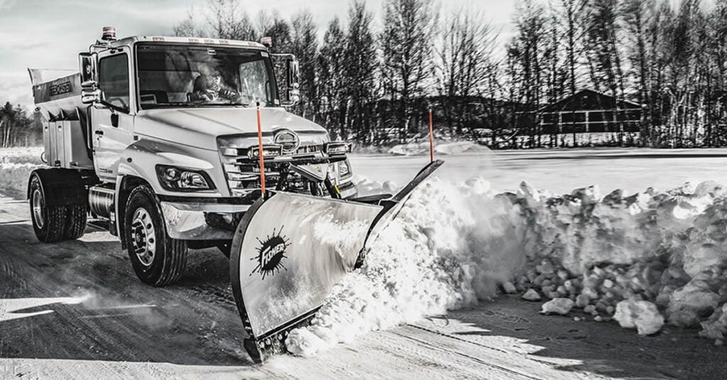 A large truck equipped with a snow plow clears snow from a road on a winter day, ensuring safe passage for food shipping vehicles amid trees, houses, and a snowy landscape in the background.
