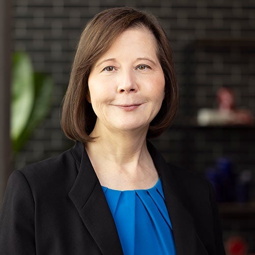 Charlene York, with straight brown hair, wears a black blazer over a blue blouse and smiles softly. She is posed in front of a blurred dark brick background with some shelves in the distance—an xModal portrait of quiet confidence.