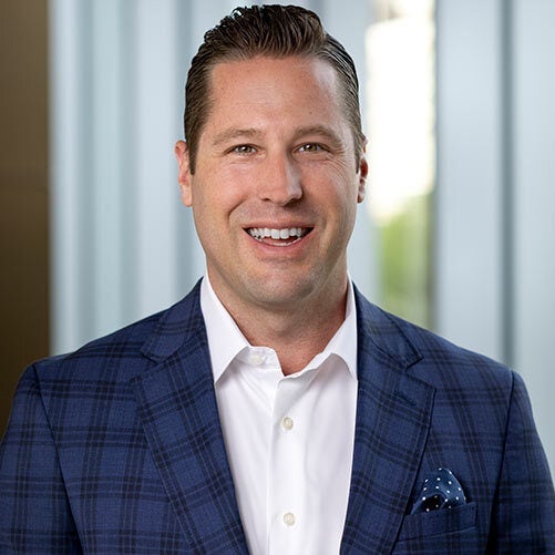 A man with short, dark hair wearing a blue plaid suit jacket, white dress shirt, and a polka dot pocket square smiles at the camera. The background features blurred vertical window panels at xModal, where JJ Lewis is often seen.