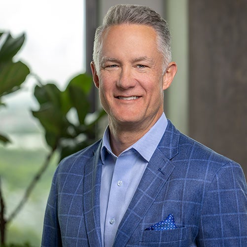 Smiling man, Joe Kniple, with short gray hair wearing a blue checkered suit jacket, light blue shirt, and blue pocket square, stands indoors in front of a window and green plant—perfect for the xModal team profile.