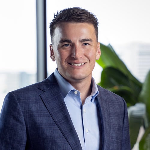 A man in a blue suit jacket and light blue dress shirt, representing a worldwide express company, smiles at the camera. He has short brown hair and stands indoors near a window with blurred city buildings and green plants behind him.