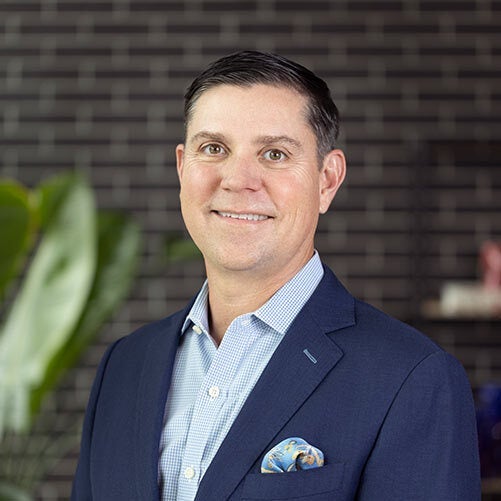 A man in a blue suit with a patterned pocket square stands in front of a dark brick wall and a large green plant, smiling confidently like the face of a worldwide express company.