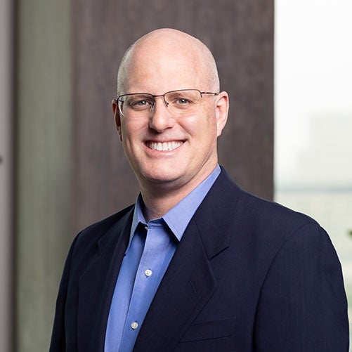 A smiling bald man, Scott Ball, wearing glasses, a blue dress shirt, and a dark blazer stands indoors near a window with blurred cityscape in the background, reflecting his role at xModal.