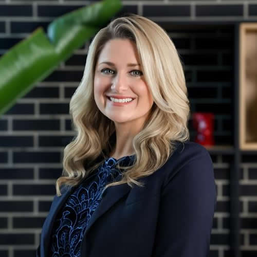 A woman with long blonde hair, wearing a dark blazer and patterned top, smiles while standing in front of a black brick wall with a green plant in the foreground at the Shelly Patman POPUP event.