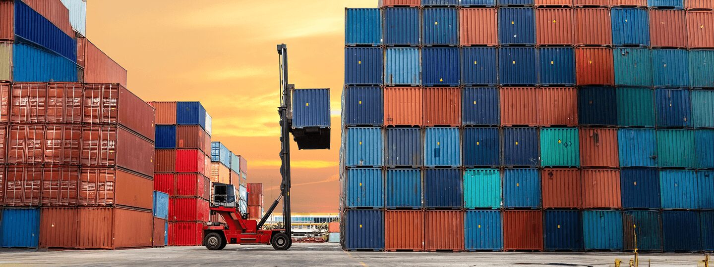 A red forklift moves a shipping container amid large stacks of colorful cargo containers at a shipping yard, where accessorial charges may apply, all set against the backdrop of an orange sunset sky.