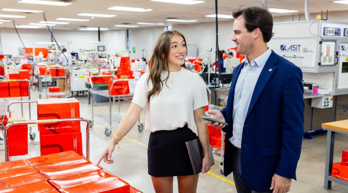 A woman and man talk in a brightly lit factory. Smiling and pointing at red boxes, the woman holds a folder while discussing Speedship, their new transportation management system. Red boxes are stacked on carts and tables throughout the workspace.