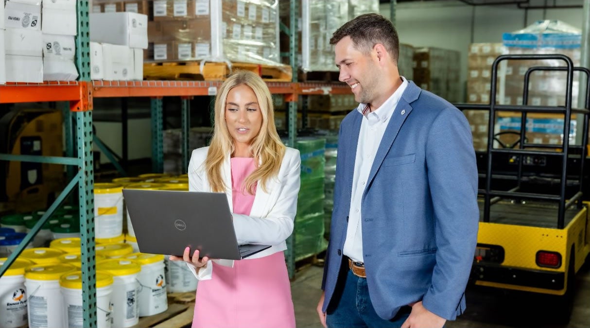 A woman in a pink dress and white blazer shows a Speedship TMS laptop screen to a man in a blue suit. They are standing and smiling in a warehouse filled with shelves, boxes, and stacked products.