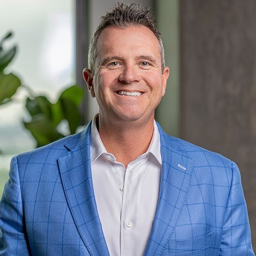 Steve Purdon, with short brown hair, wearing a light blue checkered blazer over a white shirt, smiles in a modern indoor setting with a plant and window in the background—a perfect fit for an xModal team profile.