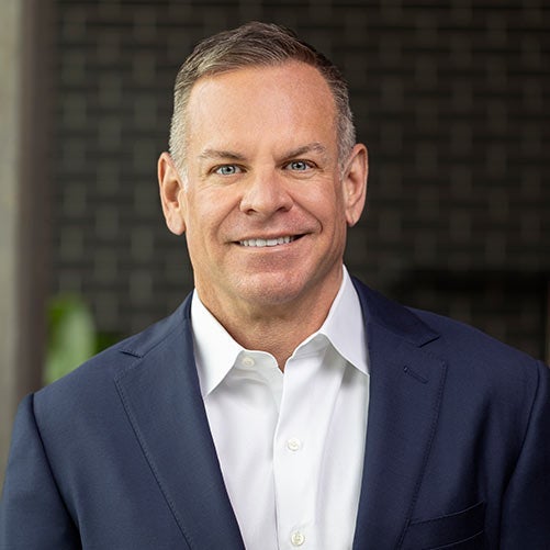 A smiling man in a navy blue suit and white dress shirt stands in front of a dark, blurred background with a subtle brick pattern, reflecting the professionalism of a worldwide express company.