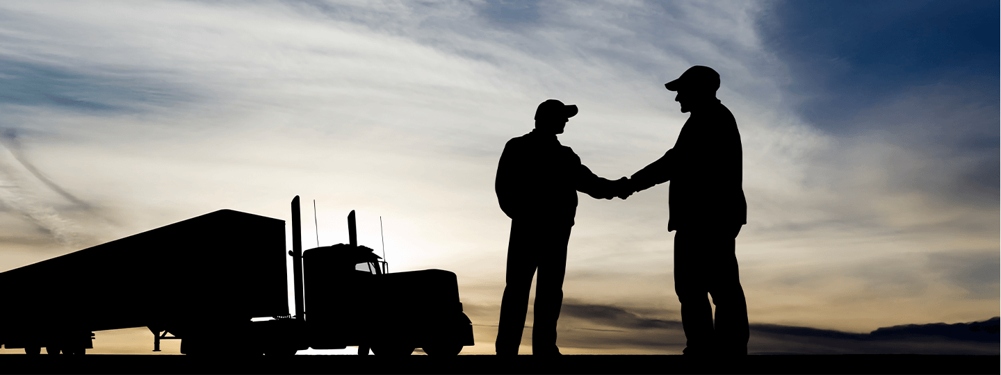 Silhouette of two people wearing hats shaking hands near a large specialty freight semi-truck, with a dramatic sky at sunset in the background.